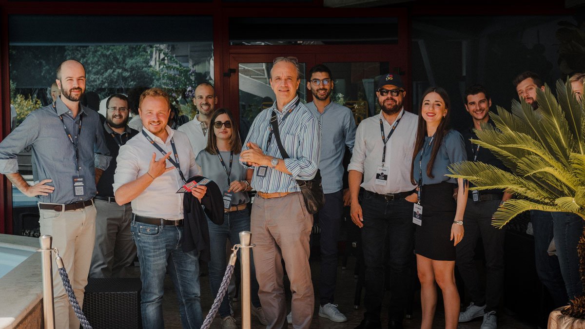 About fifteen Ennova Research team members wearing company lanyards and badges posing together outside a glass entrance on a sunny day