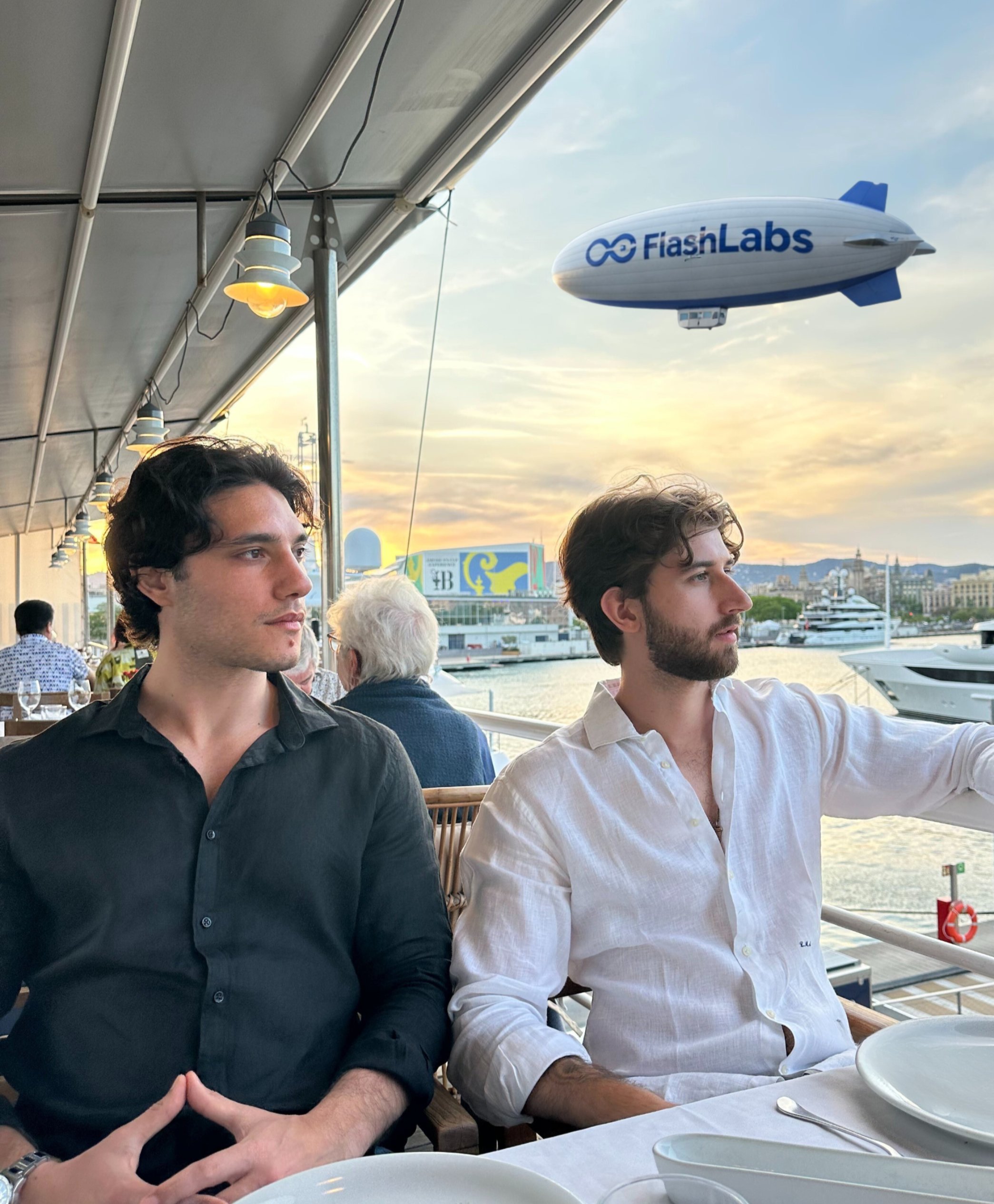 A blue and white FlashLabs blimp floats over a harbor at sunset while two men sit at a waterfront restaurant terrace below