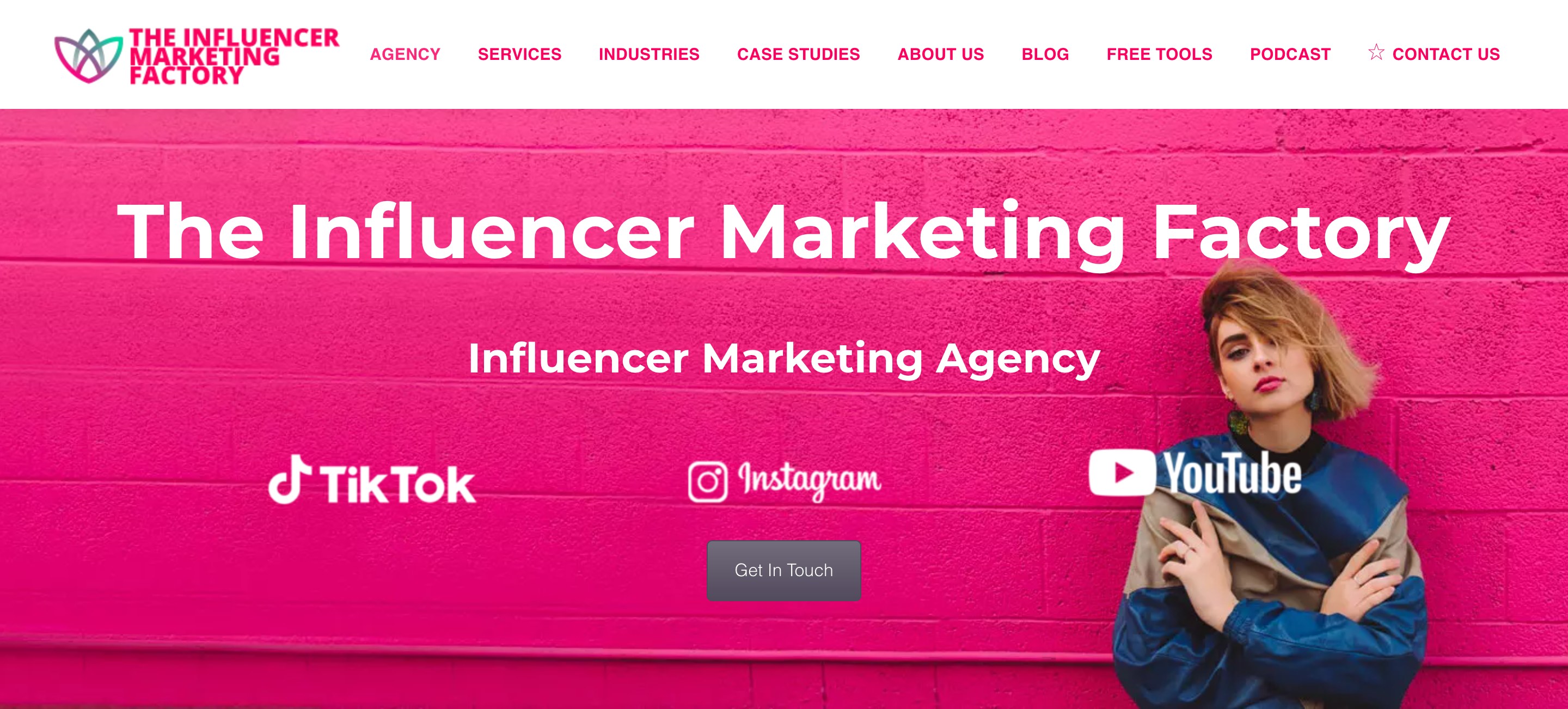 The Influencer Marketing Factory homepage: its name and the line Influencer Marketing Agency in white over a pink brick wall, the TikTok, Instagram and YouTube logos, a Get In Touch button and a woman in a blue jacket on the right
