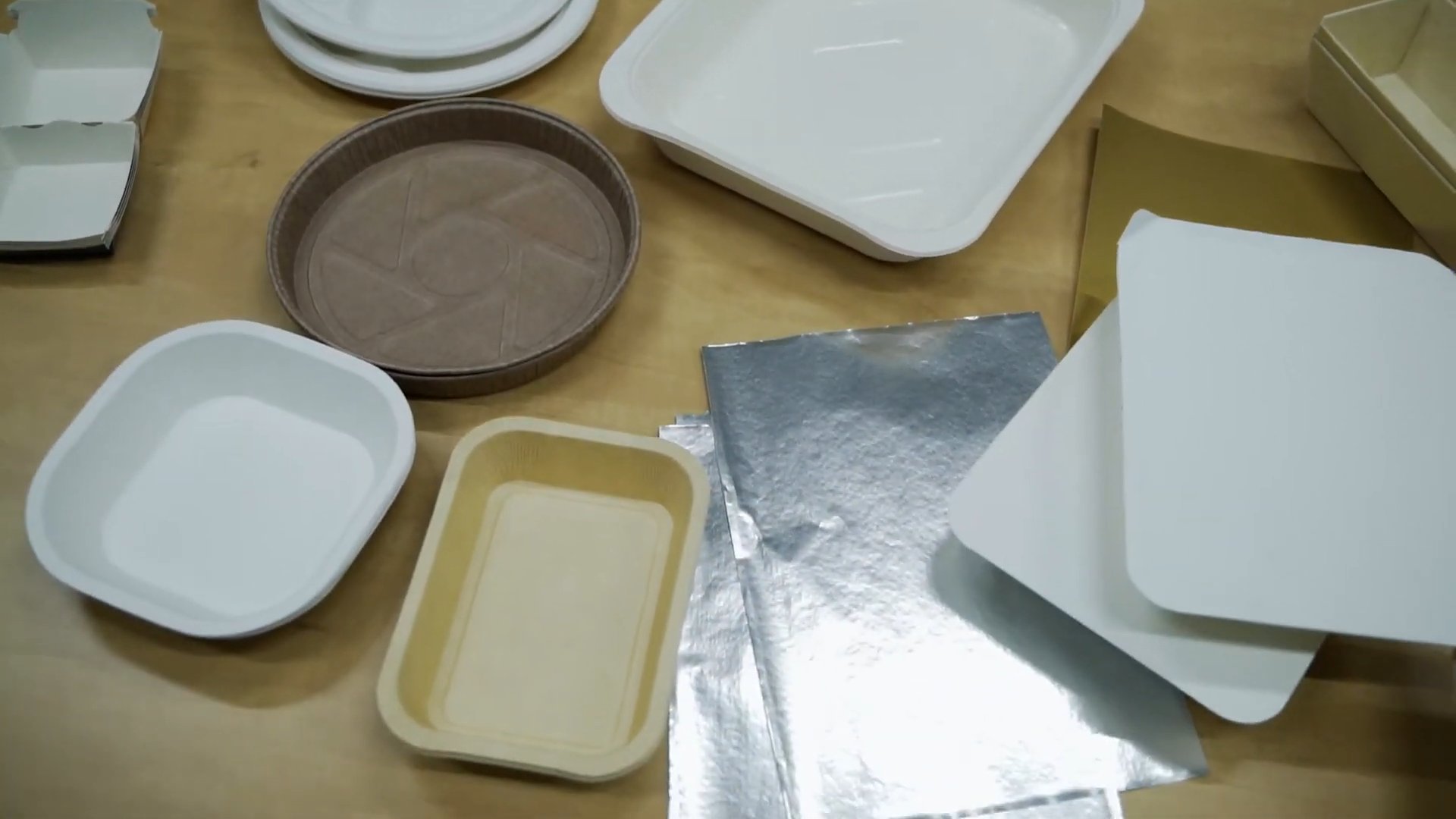 Food packaging samples on a wooden table, including molded-fiber trays, a brown paper baking tray, foil sheets and board lids
