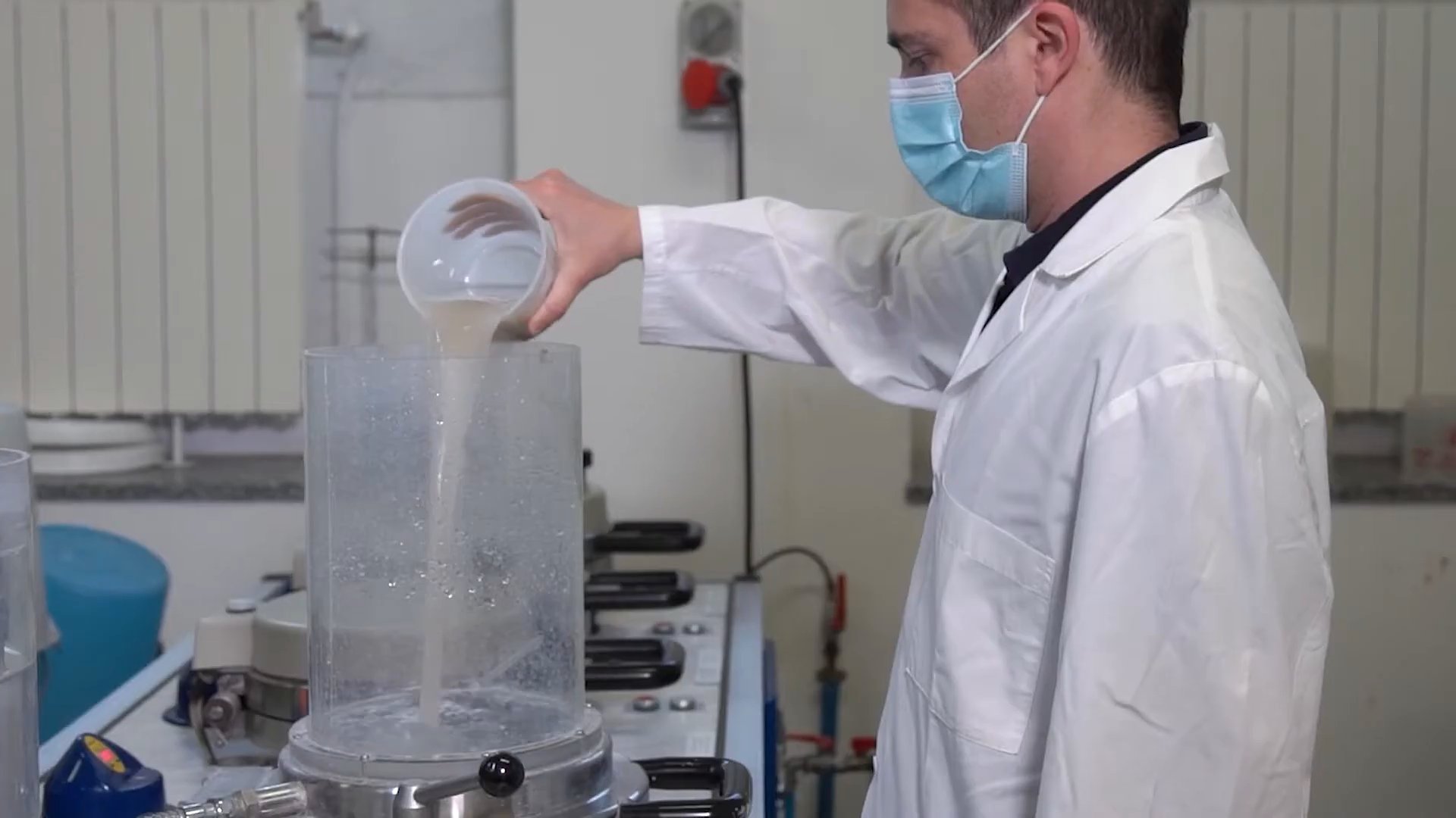 Masked technician pouring diluted pulp from a jug into the clear cylinder of a laboratory sheet former