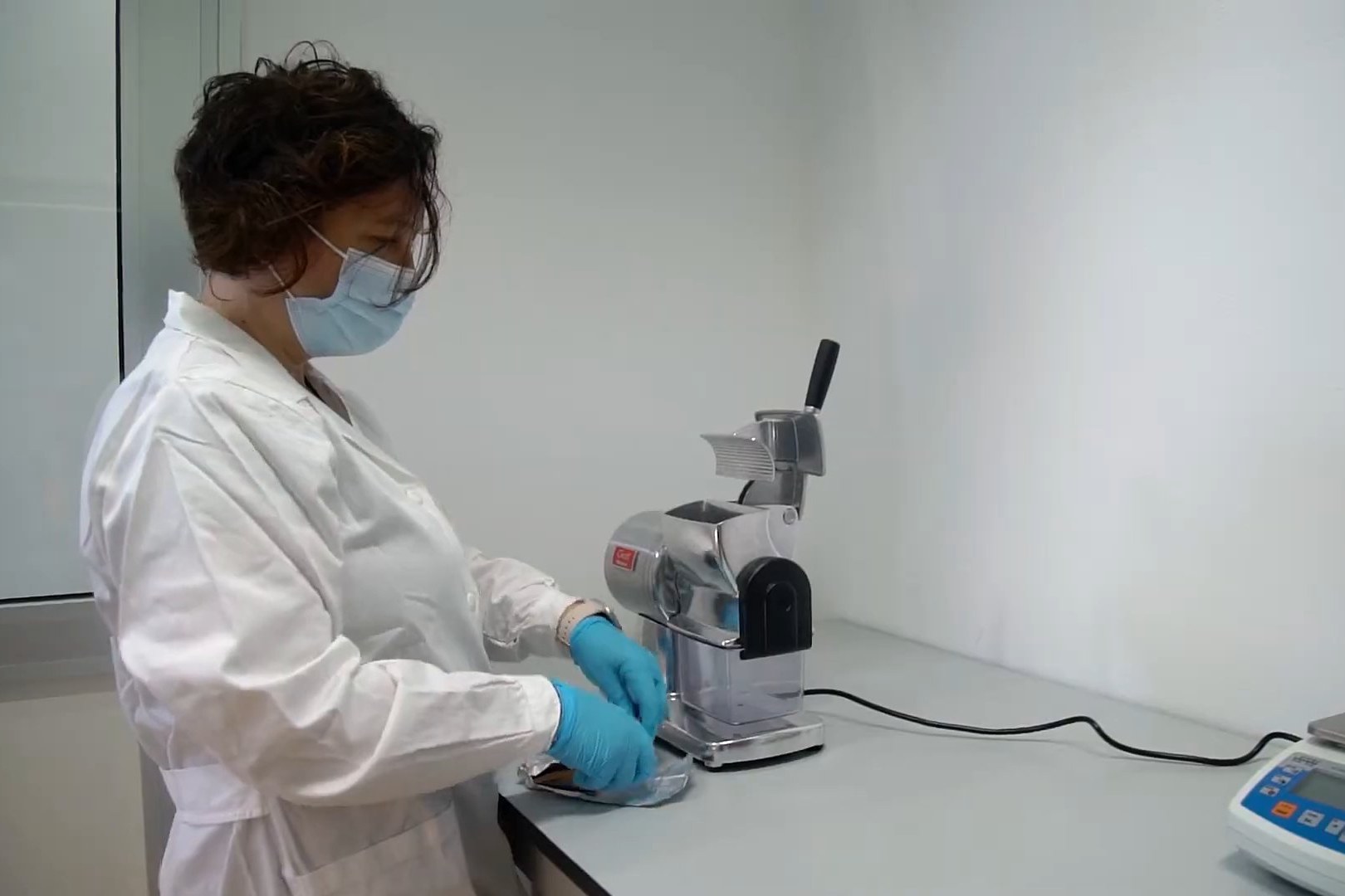 Masked lab technician in a white coat preparing a packaging sample beside a steel grinder and a digital balance