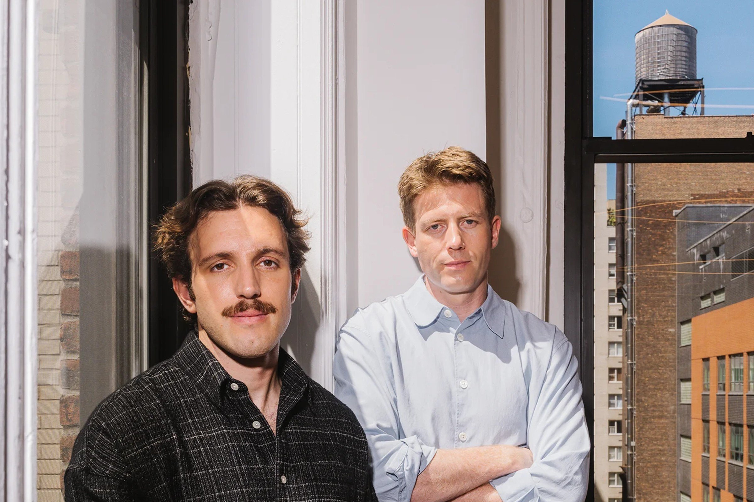 Profound co-founders Dylan Babbs and James Cadwallader standing by an office window with a rooftop water tower behind them