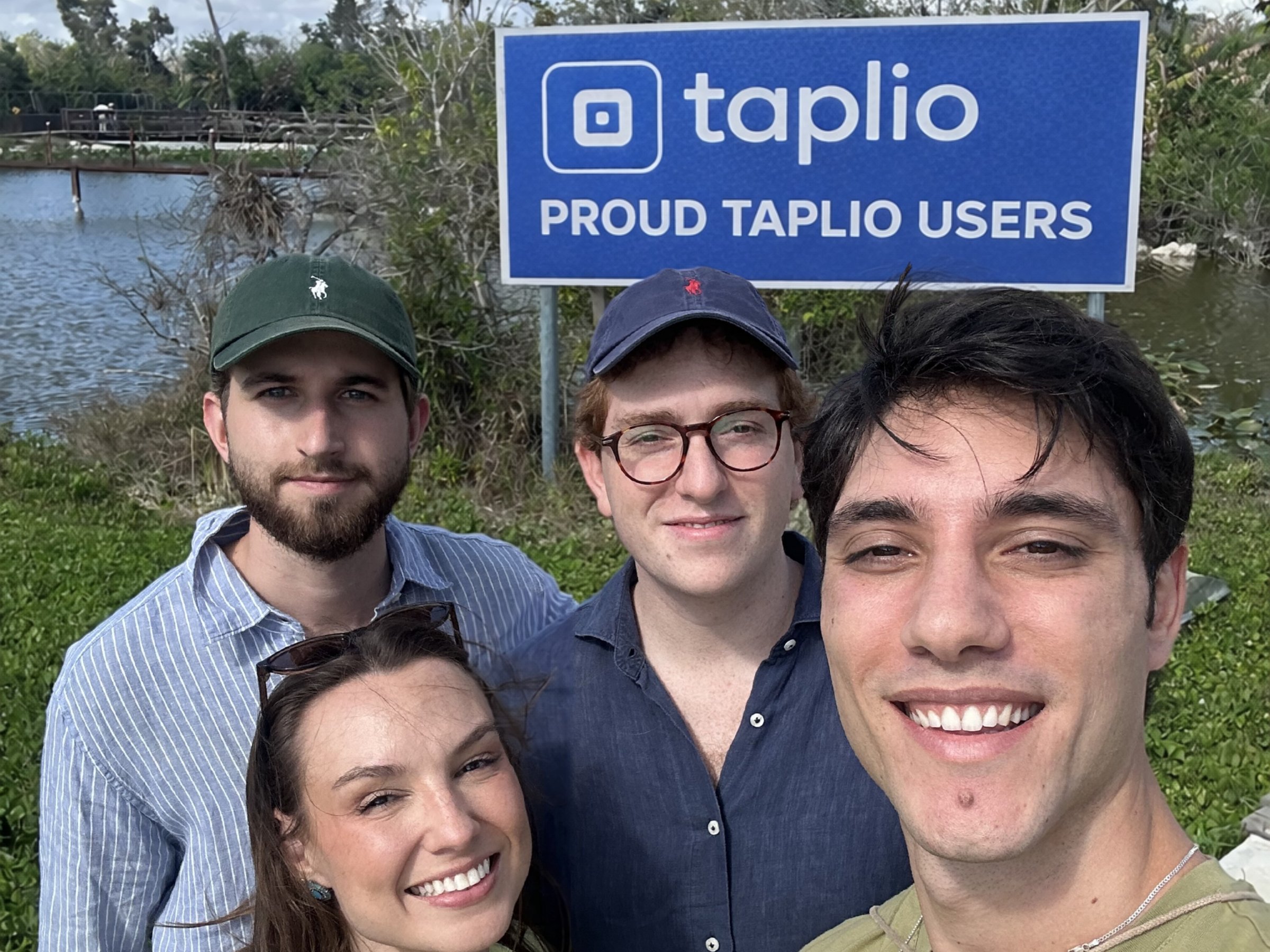Four people in a selfie in front of a blue sign that reads "taplio, Proud Taplio users" beside a lake with palm trees