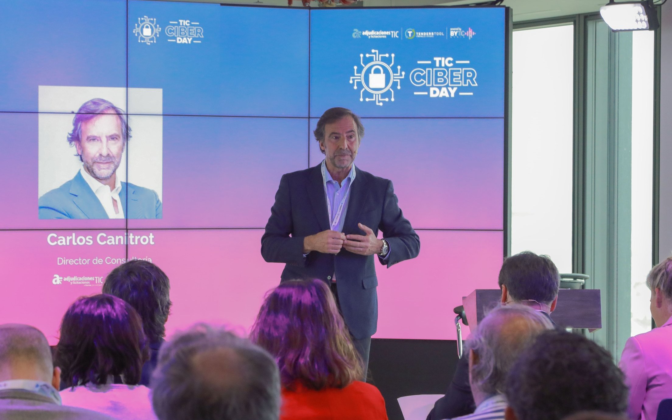 Carlos Canitrot speaking to an audience in front of a blue and pink TIC Ciber Day video wall that carries the TendersTool and adjudicaciones TIC logos
